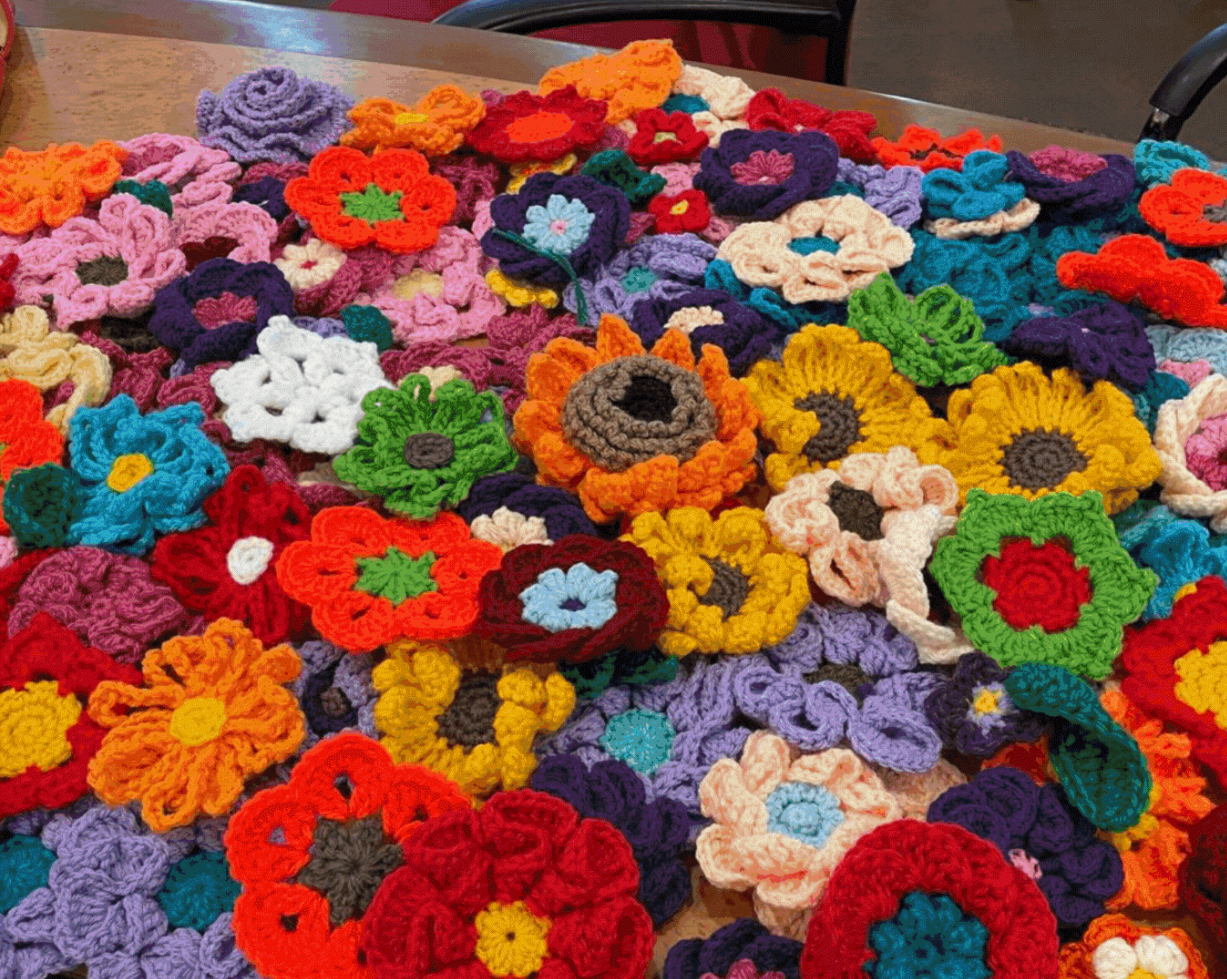 Dozens of colourful crocheted flowers in red, orange, yellow, purple, teal and white, densely arranged on a wooden table.